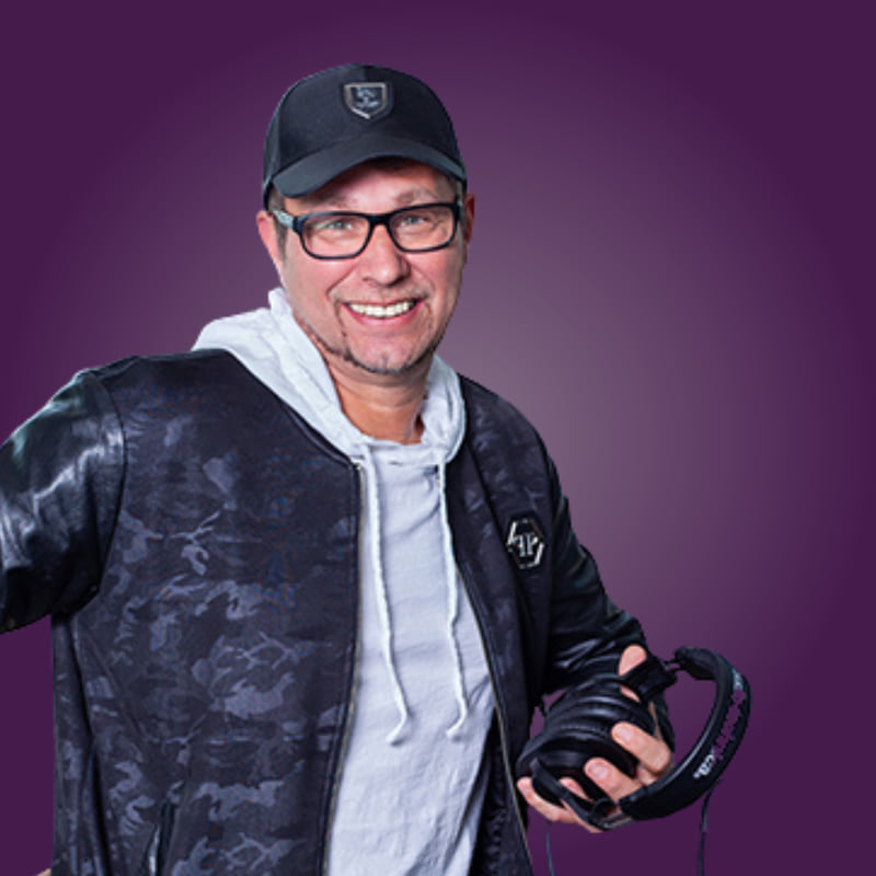 Mann mit Brille und Kappe, lächelt und hält Kopfhörer in der Hand. Hintergrund in lila Farbton.