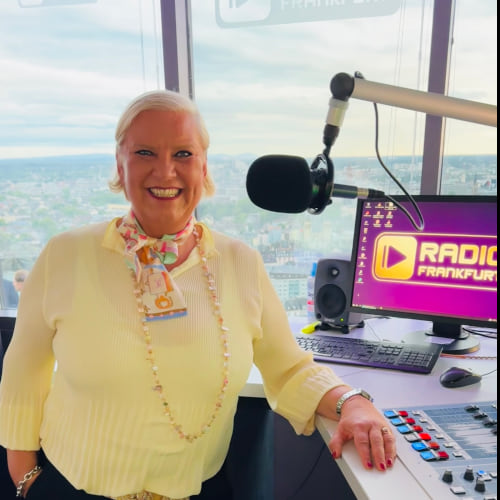 Frau mit blonden Haaren und Schal steht lächelnd vor einem Mischpult in einem Radiostudio mit Stadtansicht.