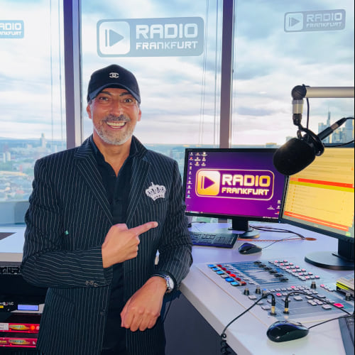 Ein Mann in gestreiftem Anzug steht an einem Radiopult mit Moderationsmikrofon und Monitoren. Im Hintergrund ist eine Stadtan