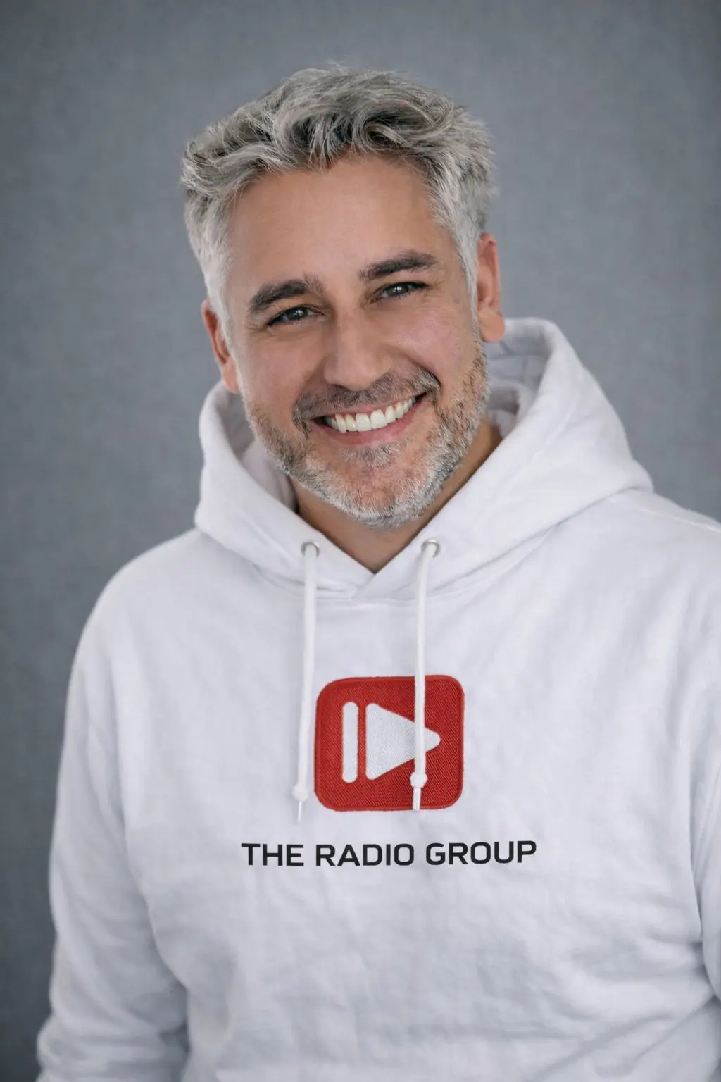 Mann mit grauen Haaren und Bart trägt einen weißen Hoodie mit rotem Logo und lächelt vor grauem Hintergrund.