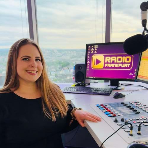 Frau mit langen Haaren sitzt an einem Radiomischpult, Monitor mit dem Logo „RADIO FRANKFURT“ im Hintergrund.
