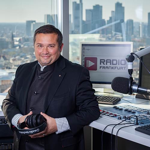Eike Knall steht in einem Radiostudio mit Blick auf die Skyline von Frankfurt und hält Kopfhörer in der Hand.
