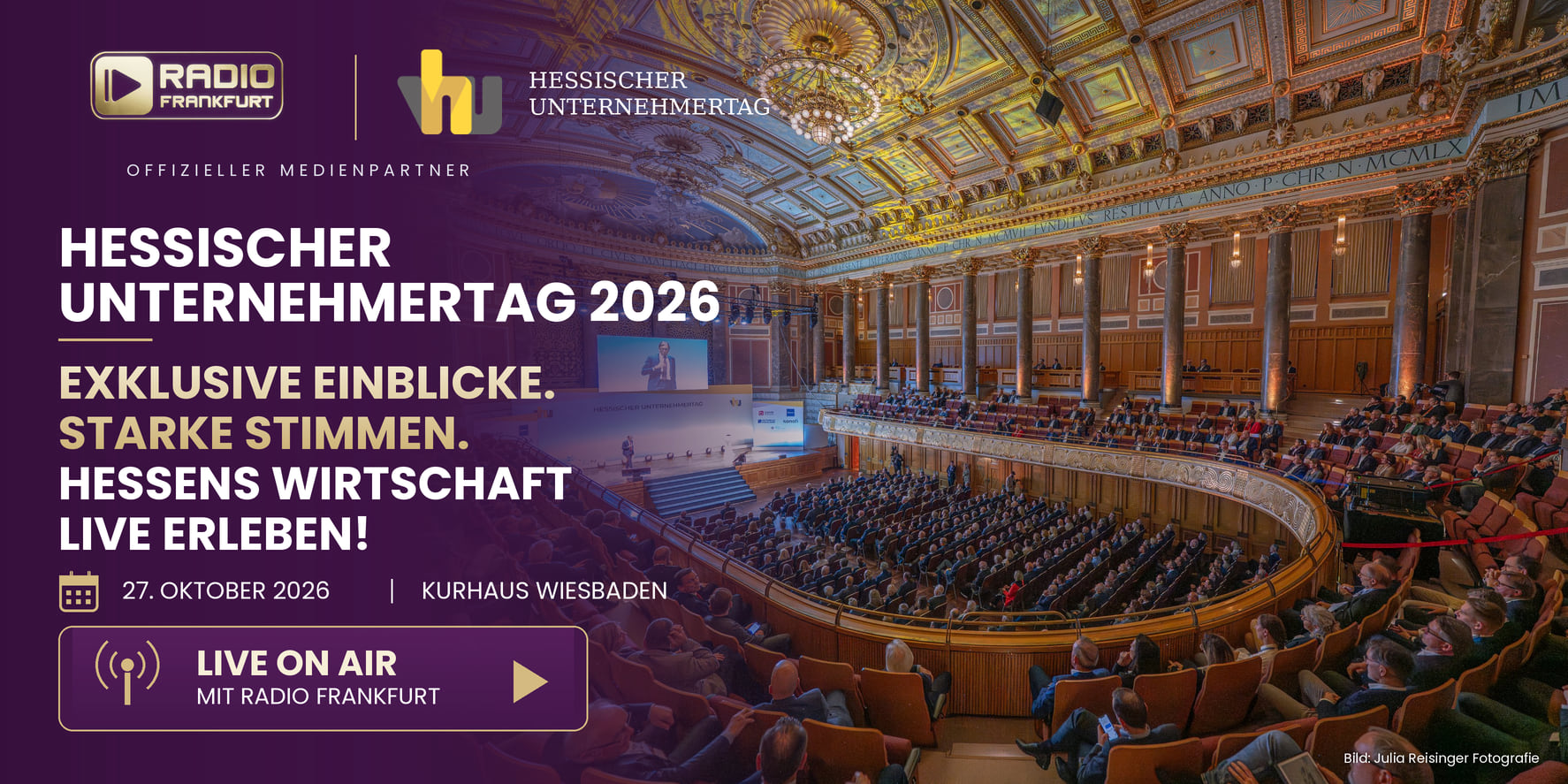 Großer Veranstaltungssaal mit Publikum. Auf der Bühne steht ein Redner. An der Wand hängen dekorative Elemente.